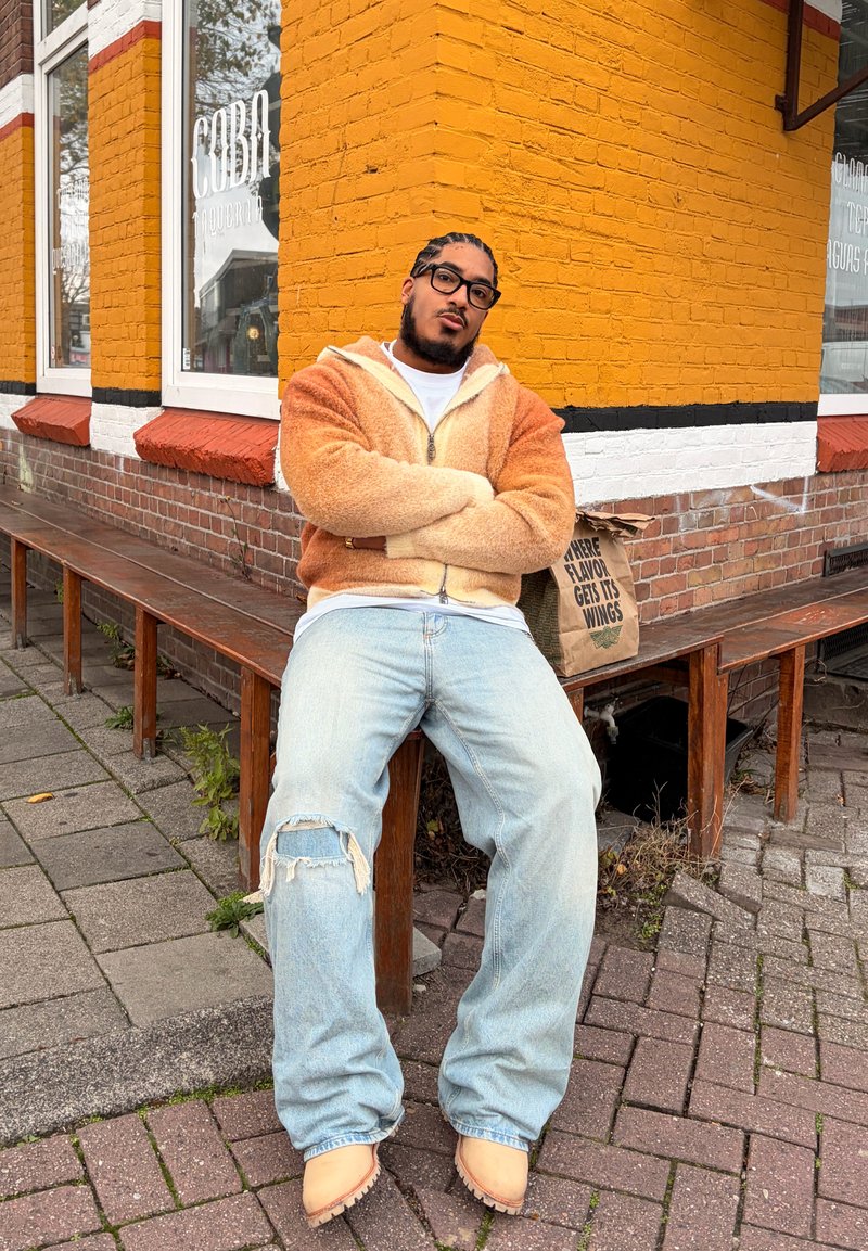 Homme portant des lunettes et des cheveux tressés assis sur un banc à l'extérieur d'un bâtiment en briques jaunes et blanches, vêtu d'une veste zippée marron et d'un jean déchiré.