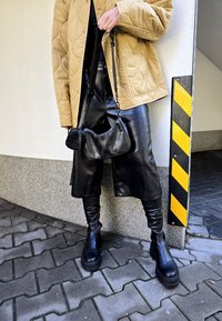 Καφέ ραμμένο μπουφάν, μαύρο δερμάτινο παντελόνι με γυαλιστερό φινίρισμα, μαύρες στρατιωτικές μπότες και μια υφασμάτινη μαύρη τσάντα με λουρί ώμου.