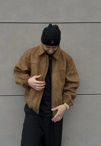 Brown suede jacket with a collared neck and elasticated cuffs, paired with a black beanie featuring a red fruit detail. Black shirt worn underneath.