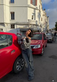 Czerwony hatchback, w paskowanym swetrze i za dużych dżinsach. Scena uliczna z pobliskimi taksówkami i białym budynkiem w tle.