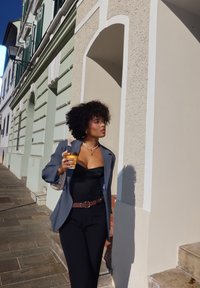 Uma pessoa vestindo um blazer cinza sobre um top de corpete preto e calças pretas segura uma caneca. O fundo apresenta um edifício de cor clara com persianas verdes.