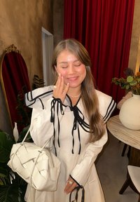 Hvid kjole med lange ærmer og sorte bånd ved kraven. Vævet hvid håndtaske og perlehalssmykke. Baggrunden har røde gardiner.