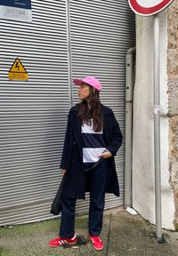 Μαύρο παλτό τρέντσι, μπλε και λευκή ριγέ μπλούζα, σκούρα τζιν, κόκκινα αθλητικά παπούτσια με λευκές ρίγες και ροζ καπέλο. Αστικοί χώροι με μεταλλικό φόντο.