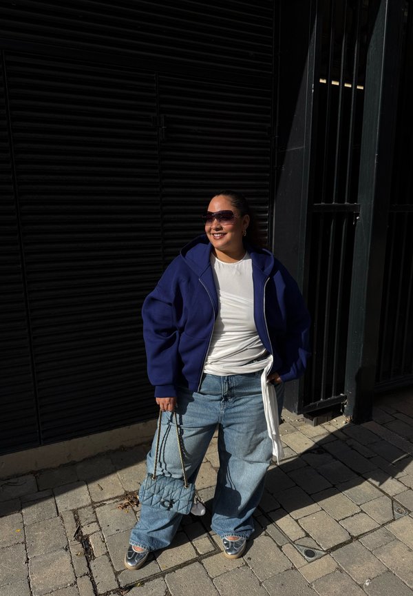 Una mujer con gafas de sol, una sudadera azul con capucha, una camiseta blanca y vaqueros holgados posa en una acera soleada mientras sostiene un bolso azul acolchado.
