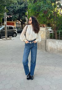 Blouse crème clair texturée avec manches bouffantes, associée à un jean en denim taille haute. Bottines noires avec détails métalliques argentés.