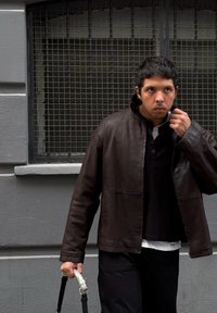 Καφέ δερμάτινο μπουφάν με ψηλό γιακά, φοριέται πάνω από ένα μαύρο πουκάμισο και λευκή μπλούζα. Κρατά ένα μπαστούνι με διακοσμητική ασημένια λαβή.