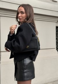 Bolso de mano de cuero negro con un diseño estructurado, acentos en metal dorado y una correa corta. Modelo llevando una chaqueta corta negra y una falda de cuero.