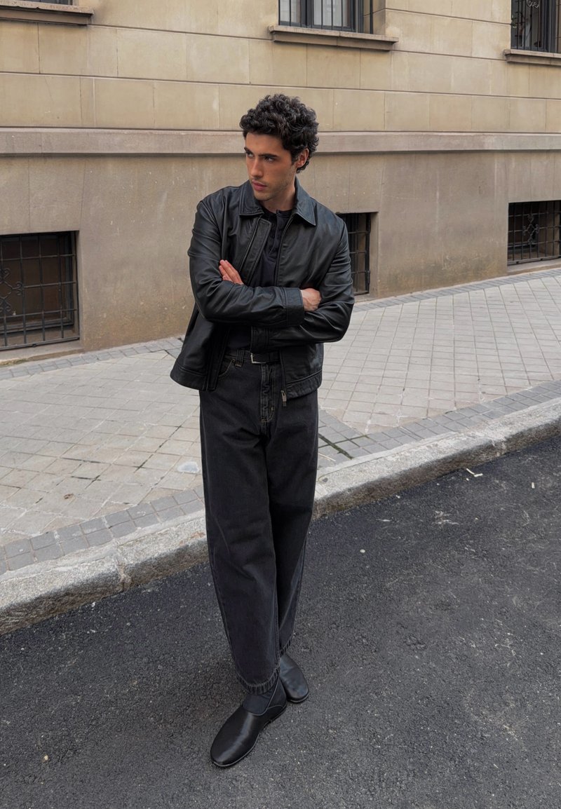 Jeune homme en veste en cuir noire, pantalon noir et chaussures noires, debout dans la rue les bras croisés, regardant vers sa gauche.