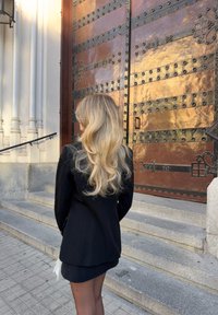 Mustmust mustriit ja seelik, õhukeste mustade sukkpükstega; pikad lainelised blondid juuksed. Taustal suured pronksist nagadega uksed ja kivised trepid.