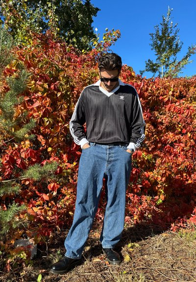 Camiseta deportiva en blanco y negro con mangas largas y escote en forma de V, combinada con unos pantalones vaqueros sueltos de color azul claro y zapatos negros. Follaje rojo de fondo.