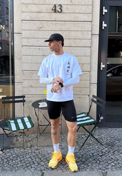 Hombre con gorra negra, camisas blancas en capas, pantalones cortos negros y zapatillas amarillas, de pie junto a una mesa y sillas de una cafetería al aire libre en el edificio número 43.