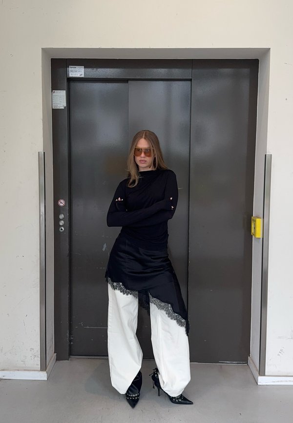 Žena u crnoj majici dugih rukava, asimetričnoj crnoj čipkanoj suknji preko bijelih širokih hlača i crnim šiljastim potpeticama stoji sa prekriženim rukama ispred zatvorenog lifta.