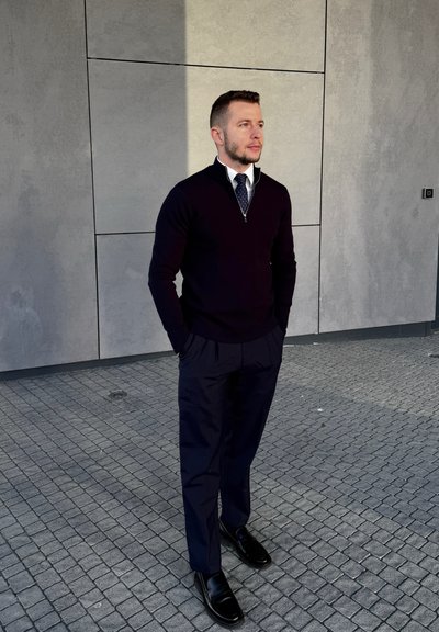 Hombre con suéter azul marino, camisa blanca, corbata de lunares y pantalones oscuros, de pie con las manos en los bolsillos sobre un pavimento de baldosas junto a una pared de paneles grises.