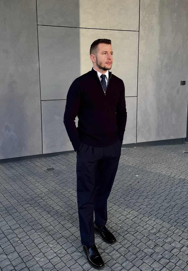 Man in navy jumper, white shirt, polka dot tie, and dark trousers stands with hands in pockets on tiled pavement by grey panel wall.