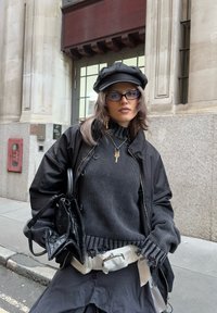 Čierna bomber bunda cez sivý pletený sveter s ošúchanými okrajmi. Doplnky zahŕňajú čiernu čiapku, okuliare a textúrovanú čiernu kabelku.