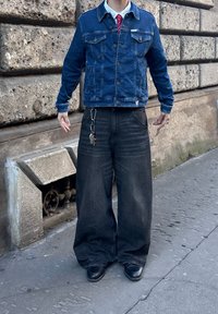 Kék farmerdzseki gombos záródással és mellzsebbel, oversize sötét farmernadrággal párosítva. Kulcstartó látható a deréknál.