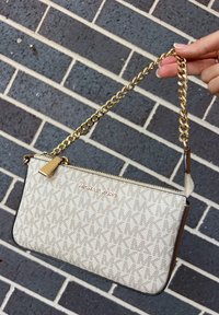 Pochette beige Michael Kors avec motif MK, fermeture éclair dorée et poignée chaîne épaisse dorée tenue par une main contre un mur de briques foncé.