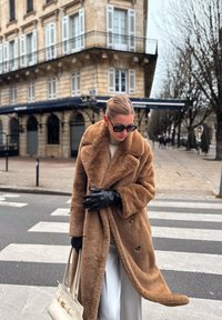 Manteau en fausse fourrure marron à double boutonnage, gants en cuir noir et sac à main de couleur claire. Trottoir et passage piéton visibles.