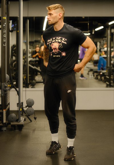 Camiseta negra de Nike con diseño gráfico, pantalones de chándal negros, zapatillas deportivas, cabello rubio corto, tatuajes, dentro de un entorno de gimnasio.