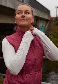 Tumesinine puffervest valge pika varrukaga topi kohal, millel on kõrge krae ja lukk. Näha on kuldsed rõngakujulised kõrvarõngad. Hall taust.