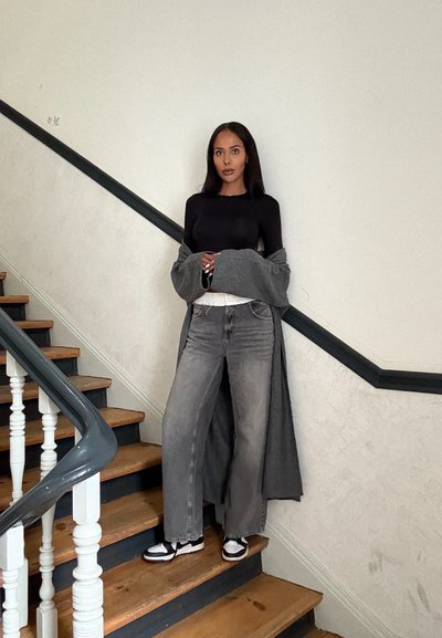 Cardigan gris sobre una camiseta negra de manga larga, combinado con jeans grises de pierna ancha y zapatos en blanco y negro, de pie en unas escaleras de madera.