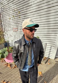 Leren jas, denim hemd, patroon stropdas en een pet met groene klep. Zonnebril op, poseert tegen een gegolfde muur en gebladerte.