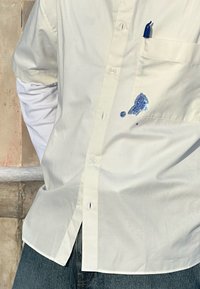 Personne portant une chemise blanche boutonnée et un jean bleu avec une tache d'encre bleue sous la poche poitrine, qui contient un stylo bleu.