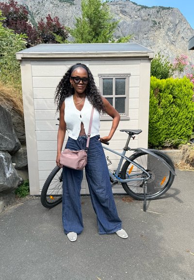 Una mujer se encuentra delante de un cobertizo de madera, vestida con un chaleco de punto blanco, jeans anchos azules y zapatos blancos con un patrón, sosteniendo un bolso cruzado rosa.