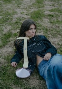 Srebrna okrągła torebka o teksturowanym wykończeniu, noszona z żółtą wstążką. Modelka ma na sobie ciemnoniebieską kurtkę dżinsową i jasnoniebieskie dżinsy.