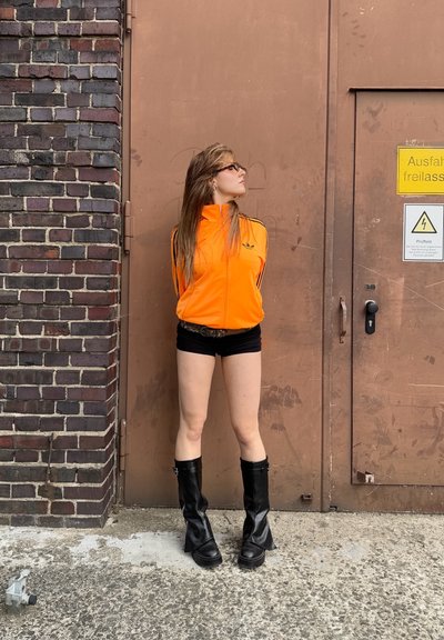 Chaqueta naranja brillante con cremallera y franjas negras, combinada con pantalones cortos negros y botas negras hasta la rodilla. El fondo presenta una pared de ladrillos y una puerta marrón.
