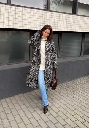 Mujer con abrigo de estampado de leopardo, suéter de cuello alto blanco, jeans azul claro, botas negras y bolso marrón, caminando por una acera de la ciudad.