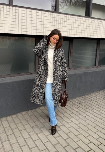 Mujer con abrigo de estampado de leopardo, suéter de cuello alto blanco, jeans azul claro, botas negras y bolso marrón, caminando por una acera de la ciudad.