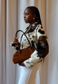 Marron sac à main en daim avec des poignées arrondies ; porté avec une veste en fausse fourrure à motifs crème et marron, et un jean blanc devant un fond neutre.