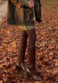 Hnědé kožené kozačky ke kolenům s naskládaným podpatkem a knoflíkovými ozdobami, v kombinaci s tmavě zeleným svetrem a černými punčochami, položené na listech.