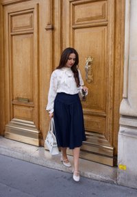 Witte blouse met een strik, donkerdenim midi-rok, witte handtas en lichtgekleurde schoenen. Houten deur op de achtergrond met gouden accenten.