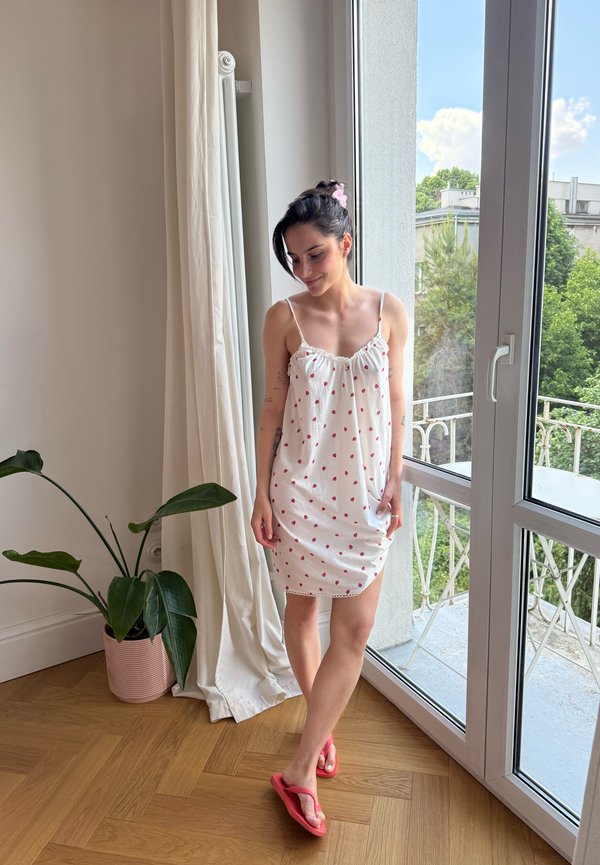 Frau in einem weißen Kleid mit roten Punkten steht barfuß neben einer Glastür, neben einer Topfpflanze und einem weißen Vorhang.