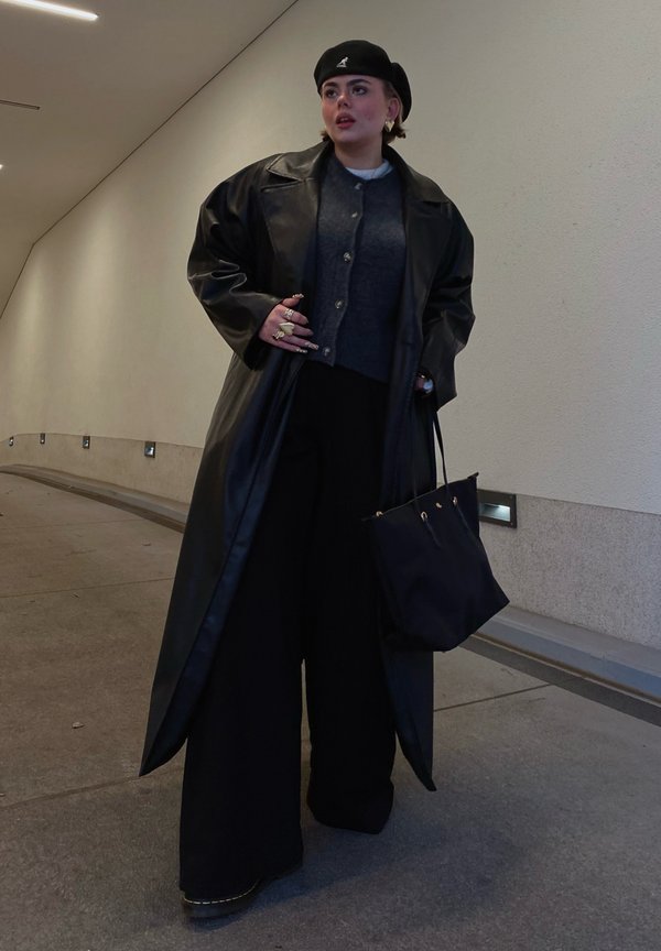 Cappotto lungo in pelle nera sopra un maglione grigio, abbinato a pantaloni neri larghi e una borsa nera. Gli accessori includono anelli dorati e un berretto nero.
