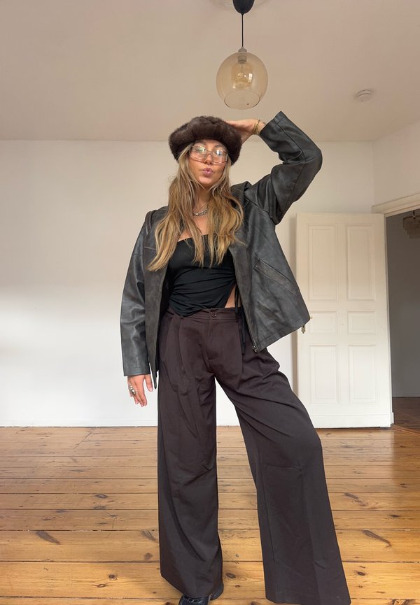 Jeune femme en intérieur portant un chapeau de fourrure marron, des lunettes, une veste en cuir noire, un haut noir et un pantalon large marron sur un sol en bois.