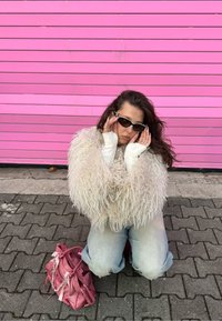 Bílý nadýchaný kožich, přiléhavá bílá košile s dlouhým rukávem, černé sluneční brýle, světle modré džíny, malá růžová kabelka na dlážděné zemi.