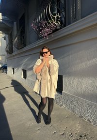 Roomkleurige cape met bontafwerking, lange mouwen en een voorkant met strik. Zwarte panty's en schoenen, met op de achtergrond een sierlijk gebouw en schaduwen.