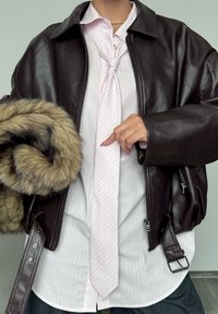 Veste bomber en cuir marron avec des zips, superposée sur une chemise rayée rose clair et une cravate nouée. Tenant un châle en fourrure.