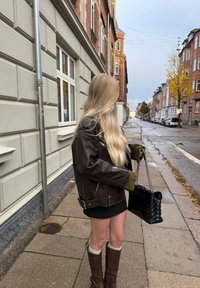 Bruine leren jas met metalen accenten, groene gebreide manchetten, zwarte mini rok en bruin kniehoge laarzen. Houdt een zwarte gewatteerde handtas vast.