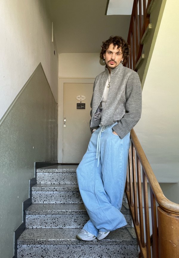 Jonge man met krullend haar leunt op de trapleuning, hij draagt een grijze jas, een licht overhemd, wijde lichtblauwe broek en witte sneakers binnen.