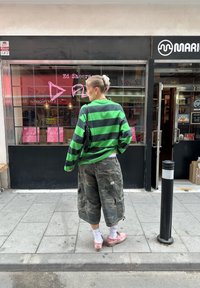 Grün-schwarz gestreiftes, �übergroßes Langarmhemd, Camouflage-Cargo-Shorts, pinke Schuhe, weiße Socken, schwarze Tasche, städtische Umgebung.