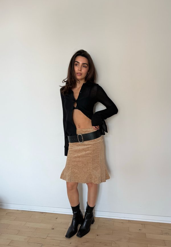 Femme debout contre un mur uni portant un cardigan court noir, une jupe beige avec une large ceinture noire, et des bottines noires.
