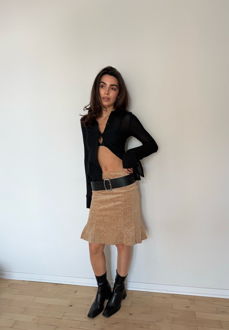 Femme debout contre un mur uni portant un cardigan court noir, une jupe beige avec une large ceinture noire, et des bottines noires.