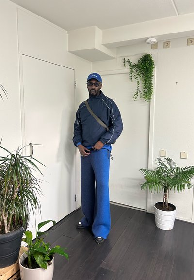 Pantalones de forro polar azules y una chaqueta azul oscura con ribete blanco. Gafas de sol negras y una gorra azul. Entorno interior con plantas en macetas.