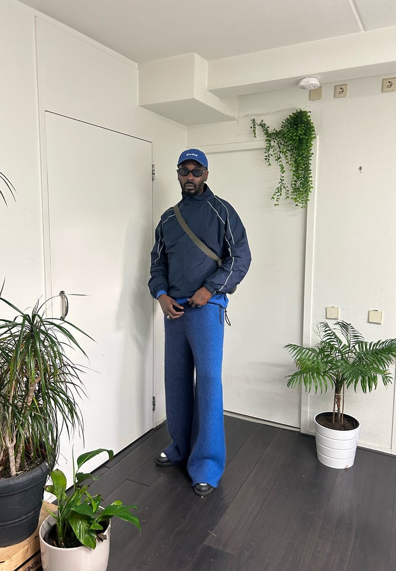 Blaue Fleecehosen und eine dunkelblaue Jacke mit weißen Akzenten. Schwarze Sonnenbrille und eine blaue Mütze. Inneneinstellung mit Pflanzen in Töpfen.
