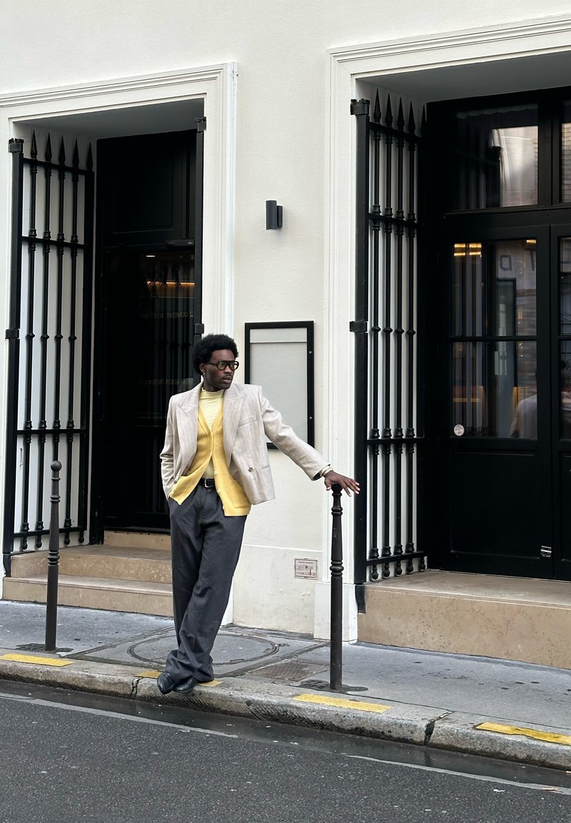 Hombre con gafas y cárdigan amarillo apoyado en un poste negro en la acera frente a un edificio blanco con puertas negras y rejas de hierro.