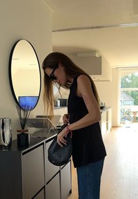 Črna tkanina torba, ki jo drži oseba v črnem brezrokavniku in modrih kavbojk, v svetli notranjosti z okroglim ogledalom in dekoracijo.